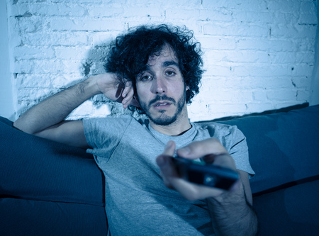 Young bored man on couch using TV remote control zapping for another movie or show late at night. Looking disinterested and sleepless. In entertainment People insomnia and Sedentary lifestyle concept.の写真素材