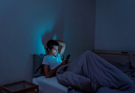Addicted man chatting and surfing on the Internet with smart phone late at night in bed. Bored,...