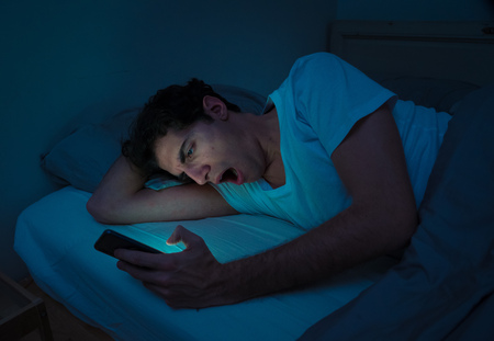 Addicted man chatting and surfing on the Internet with smart phone late at night in bed. Bored, sleepless and tired in dark room with moody light. In insomnia and mobile addiction concept.の写真素材