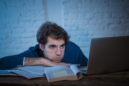 Overworked and tired male student working late at night on laptop trying not to fall asleep feeling fatigued, worried and sad. Moody dark light. Finals exams Online learning and overwork concept.の写真素材