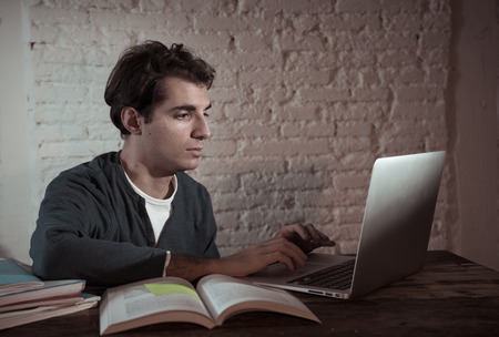 Close up portrait in moody light of happy casual attractive man working and studying on laptop sitting on desk at night. In working from home, online learning and final exams concept.の写真素材