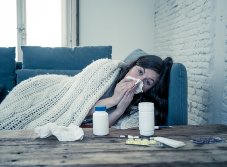 Sick woman on sofa sneezing and blowing her nose with allergy, hay fever or cold feeling unwell, fatigue and restless not being able to go to work. Lifestyle portrait In Health Care and medical.の写真素材