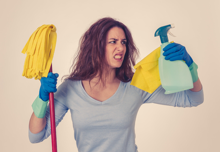 Beautiful angry and upset woman holding mop and cleaning spray feeling frustrated and furious in domestic duties concept and gender stereotypes. Studio portrait isolated on blue background.の写真素材