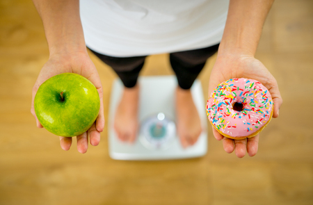 Close up of woman on scale holding on hands apple and doughnut making choice between healthy unhealthy food dessert while measuring body weight in Nutrition Health care Diet and temptation concept.の写真素材