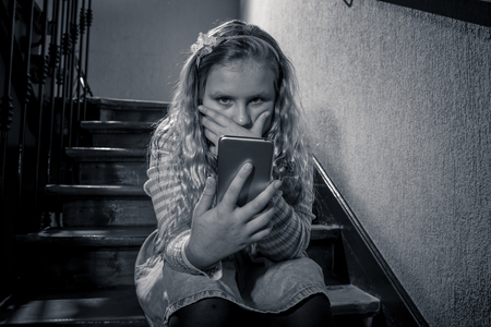 Sad depressed young girl victim of cyberbullying by mobile smart phone sitting on stairs feeling lonely, unhappy, hopeless and abused. Child bullied and harassed by text message by online stalker.の写真素材