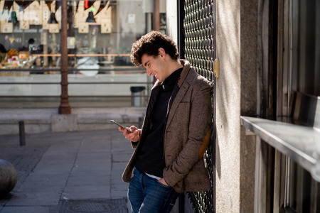 Fashion college man in his twenties happy checking blog or chatting on the internet with friend on smart mobile phone app in european city outdoors city. In communication technology and travel Europe.の写真素材