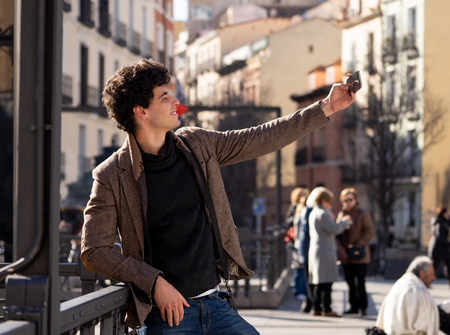 Excited trendy young man taking selfie or blogging on smart mobile phone on holidays in european city street or university campus. Holidays and technology Connection and social media concept.の写真素材