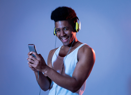 Strong african american young man listening to music in smartphone app looking for best song online with headphones singing and dancing. Portrait with disco dance floor light in Music and technology.の写真素材