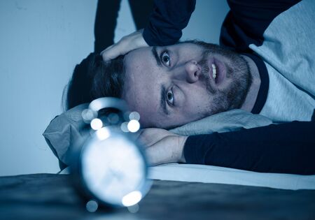 Sleepless and desperate young caucasian man awake at night not able to sleep, feeling frustrated and worried looking at clock suffering from insomnia in stress and sleeping disorder concept.の写真素材
