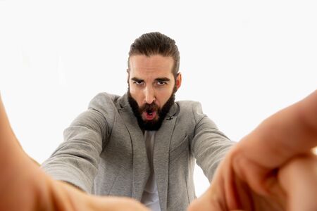 Young funny bearded hipster blogger man in modern casual style holding camera or smart phone shooting self portrait or recording video for his followers. In internet social media, blog concept.の写真素材