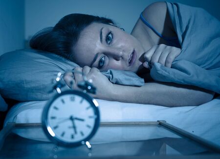 Sleepless and desperate beautiful caucasian woman awake at night not able to sleep, feeling frustrated and worried looking at clock suffering from insomnia in sleep disorder concept.の写真素材
