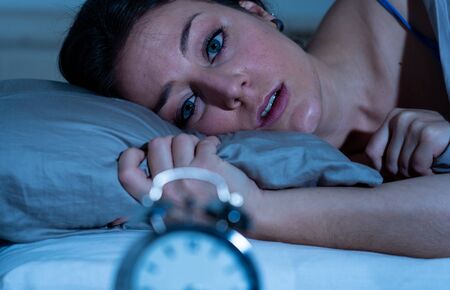 Sleepless and desperate beautiful caucasian woman awake at night not able to sleep, feeling frustrated and worried looking at clock suffering from insomnia in sleep disorder concept.の写真素材