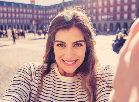 Beautiful young tourist student happy and excited in Plaza Mayor Madrid holding the camera and taking a selfie in plaza Mayor Madrid Spain. In tourism, European city, travel in Europe and connections.の写真素材