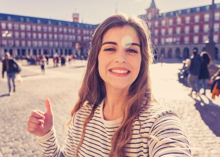Beautiful young student tourist woman happy and excited in Plaza Mayor Madrid taking a selfie holding the mobile or tablet. Looking cheerful making thumbs up gesture. In tourism and travel in Europe.の写真素材