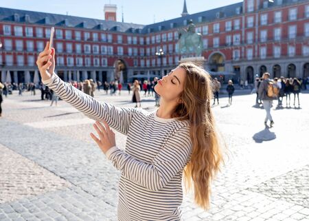 Beautiful young caucasian student tourist woman taking a selfie or video for her tourism and travel around the world blog web in Plaza Mayor Madrid Spain. In social media and vacations in europe.の写真素材