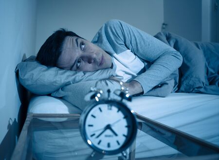 Sleepless and desperate young caucasian man awake at night not able to sleep, feeling frustrated and worried looking at clock suffering from insomnia in stress and sleep disorder concept.の写真素材