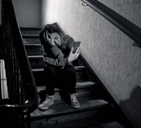 Sad depressed young teenager girl victim of cyberbullying by mobile smart phone siting on stairs feeling lonely, unhappy, hopeless and abused. Bullied by text message on social media app. Dark lightの写真素材