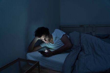 Addicted man chatting and surfing on the Internet with smart phone late at night in bed. Bored, sleepless and tired in dark room with moody light. In insomnia and mobile addiction concept.の写真素材
