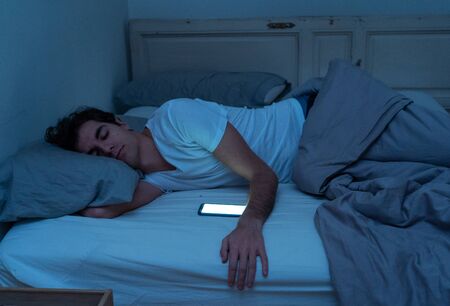 Addicted to social media young man falling asleep with smart mobile phone at night in bed. lifestyle portrait of man sleeping in dark bedroom with mobile screen light on. Mobile use addiction.の写真素材