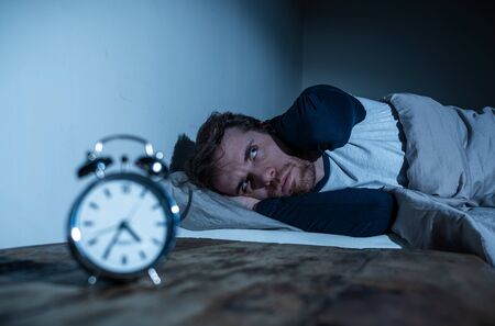 Insomnia Stress and Sleeping disorder concept. Sleepless desperate young caucasian man awake at night not able to sleep, feeling frustrated and worried looking stressed and concerned at alarm clock.の写真素材