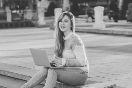 Pretty young cheerful teenager woman working or studying on laptop computer sitting happy and on stairs in city outdoors urban background. In student lifestyle Online education and Technology concept.の写真素材