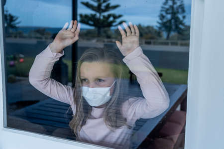 COVID-19 Lockdown. Depressed lonely little girl with face mask looking through the window during quarantine. Sad sick child in self isolation at home. Coronavirusu outbreak and children mental health.の写真素材