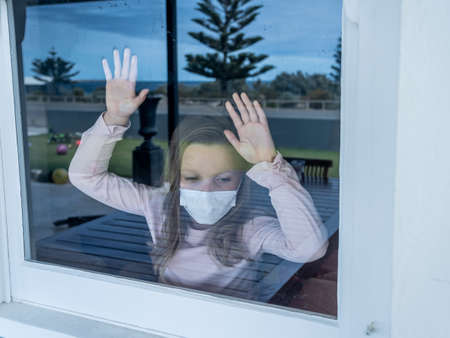 COVID-19 Lockdown. Depressed lonely little girl with face mask looking through the window during quarantine. Sad sick child in self isolation at home. Coronavirusu outbreak and children mental health.の写真素材