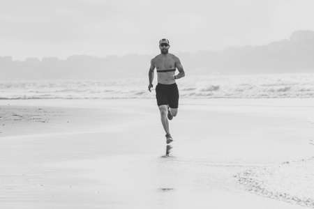 Sports fitness man with heart rate monitor running on beach with smart watch. Fit body male runner doing jogging workout at the sea in sport exercise and technology and healthy lifestyle concept.の写真素材