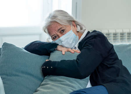Lonely depressed senior widow woman with protective mask crying on couch isolated at home, sad and worried missing husband and family in COVID-19 death, lockdown, social distancing and Mental health.の写真素材