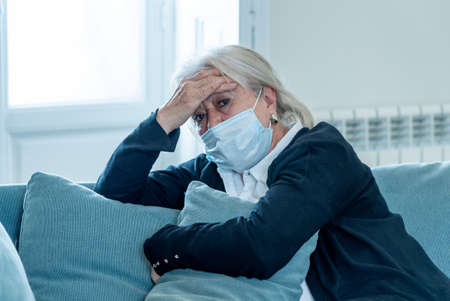 Lonely depressed senior widow woman with protective mask crying on couch isolated at home, sad and worried missing husband and family in COVID-19 death, lockdown, social distancing and Mental health.の写真素材