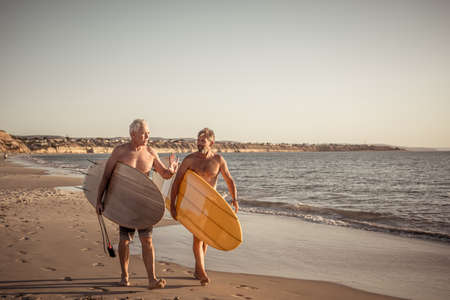 Two mature men walking with surfboards on beautiful beach enjoying paradise and retirement...