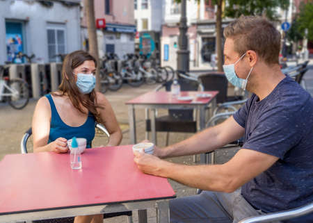 Couple sitting in a coffee shop wearing surgical protective face mask spending time outdoors in...