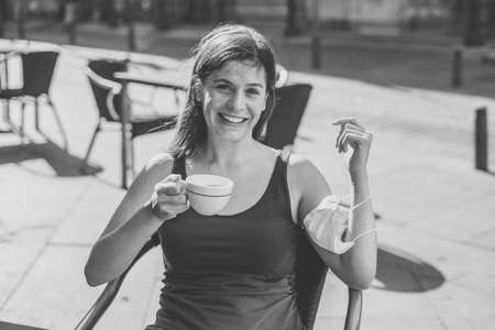 Young woman sitting in a coffee shop wearing surgical protective face mask in her arm while drinking coffee outdoors in public settings in the Coronavirus New Normal. COVID-19 Outbreakの写真素材