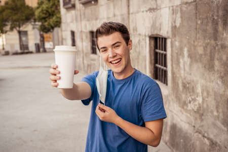 COVID-19 outbreak. Young man walking in city street wearing protective surgical face mask and drinking coffee. Coronavirus, the New Normal and mandatory use of face maskの写真素材