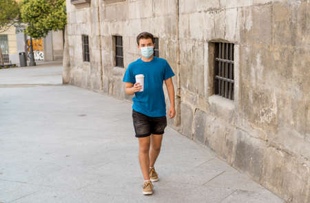 COVID-19 outbreak. Young man walking in city street wearing protective surgical face mask and drinking coffee. Coronavirus, the New Normal and mandatory use of face maskの写真素材