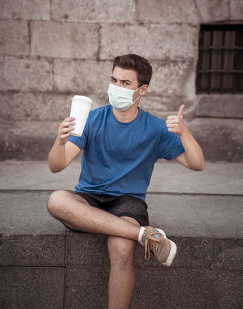 COVID-19 outbreak. Young man walking in city street wearing protective surgical face mask and drinking coffee. Coronavirus, the New Normal and mandatory use of face maskの写真素材