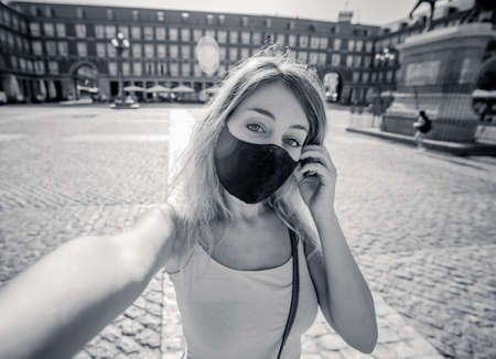 Beautiful young caucasian tourist woman wearing protective face mask happy in Plaza Mayor Madrid taking a selfie, sending a virtual kiss and showing thumbs up. COVID-19 and travel in the New Normal.の写真素材