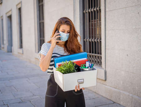 Sad businesswoman in face mask and box with office staff on mobile calling family after being fired....