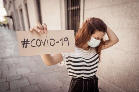 Portrait of sad depressed woman in medical protective face mask holding COVID-19 sign. Coronavirus...