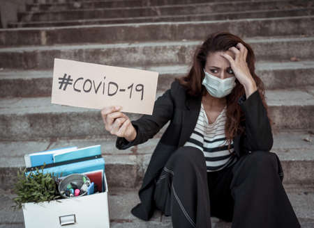 Sad depressed woman in face mask holding box with personal staff and COVID-19 sign outside office. Female worker lost her job due to coronavirus Outbreak impact in economy, business and unemployment.の写真素材