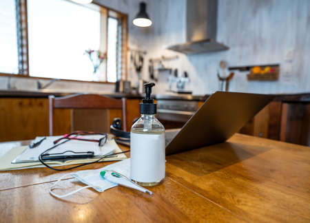COVID-19 and working from home concept. Conceptual image with no people of laptop, hand sanitizer, thermometer, surgical mask and paperwork on kitchen table as workspace in coronavirus and new normal.の写真素材