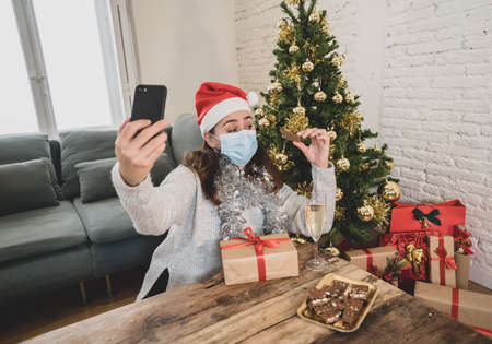 Happy woman with face mask home alone in lockdown using smart mobile cell phone, celebrating Virtual christmas video calling family and friends. COVID-19 Virtual holidays and online social gathering.の写真素材