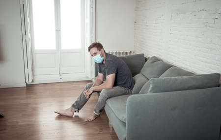 Sad man with protective face mask at home living room couch feeling tired and worried suffering depression amid lockdown and social distancing. Mental Health and isolation concept.の写真素材