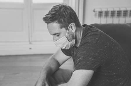 Sad man with protective face mask at home living room couch feeling tired and worried suffering depression amid  lockdown and social distancing. Mental Health and isolation concept.の写真素材