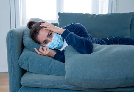 Sad latin woman with protective face mask at home living room couch feeling tired and worried suffering depression amid  lockdown and social distancing. Mental Health and isolation concept.の写真素材