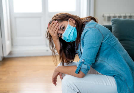 Sad latin woman with protective face mask at home living room couch feeling tired and worried suffering depression amid  lockdown and social distancing. Mental Health and isolation concept.の写真素材