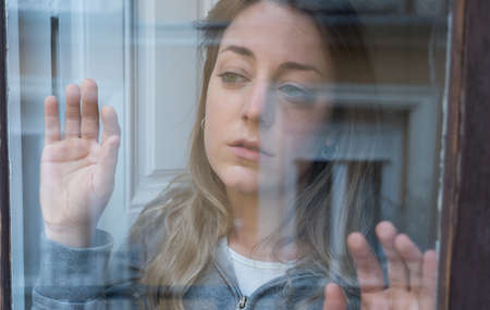 Young beautiful depressed unhappy caucasian woman looking worried and sad through the window at...