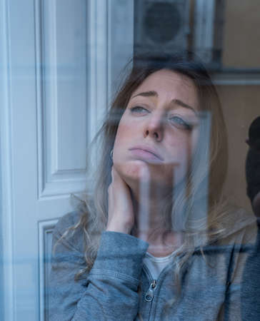Young beautiful depressed unhappy caucasian woman looking worried and sad through the window at home. Feeling worthless and in pain. closeup. Depression and Mental Health conceptの写真素材
