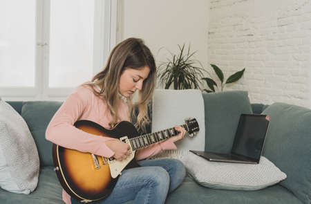 Attractive musician woman playing electric guitar during online concert at home in isolation. Music teacher teaching playing instrument online using laptop, streaming and recording courses.の写真素材