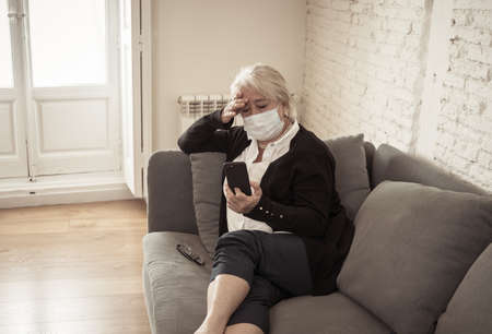 Lonely sad senior woman in face mask feeling scared, hopeless and depressed, worried about husband at hospital infected with coronavirus .Checking mobile phone for news. Anxiety, fear and COVID-19.の写真素材